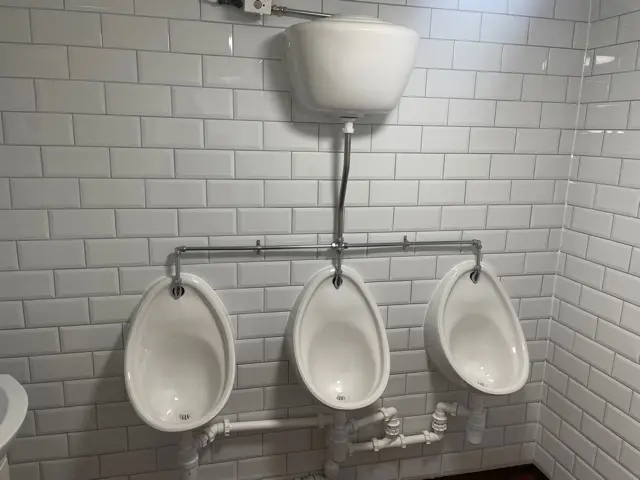 Three wall-mounted urinals beneath a high-level cistern, against white tiled walls