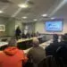 Engineers seated around tables during a team meeting, with a presentation on screen.